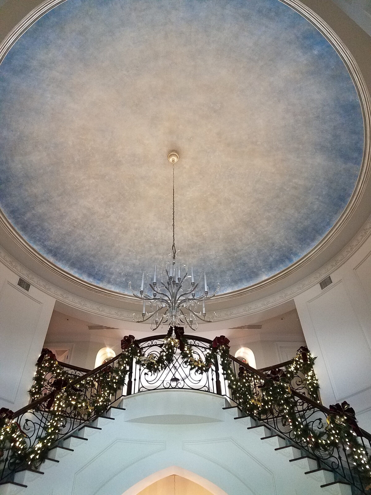 Crystal chandelier hanging from a painted dome ceiling above a double staircase